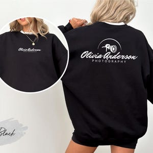 May include: Black sweatshirt with the text "Olivia Anderson Photography" in white script on the front and back. The back design includes a camera graphic. The sweatshirt has a crew neck and long sleeves.