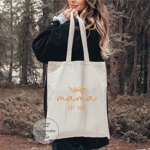 May include: A white canvas tote bag with the word "mama" in orange script with a small floral design above the word. The text "EST. 2023" is below the word "mama". A small circle with the text "choose your design color & year" is in the bottom left corner of the bag.