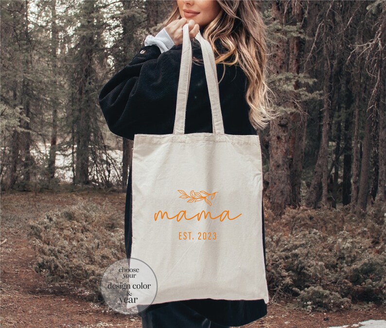May include: A white canvas tote bag with the word "mama" in orange script with a small floral design above the word. The text "EST. 2023" is below the word "mama". A small circle with the text "choose your design color & year" is in the bottom left corner of the bag.