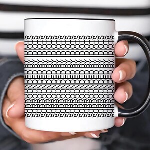 Pode incluir: Caneca de cerâmica branca com alça e borda pretas. A caneca apresenta um padrão repetitivo de desenhos geométricos pretos. A caneca é segurada por uma pessoa.