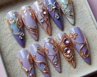 May include: A set of ten long, almond-shaped artificial nails. Each nail is painted in shades of lavender, lilac, and peach, and adorned with gold filigree and sparkling rhinestones. Some nails feature iridescent accents, creating a glamorous, ornate design.