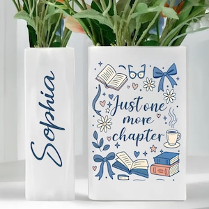 May include: Two white rectangular vases with floral arrangements. One vase has the name "Sophia" in blue script. The other vase features a blue and white illustration with the text "Just one more chapter" and images of books, glasses, and flowers.