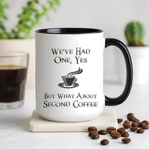May include: White coffee mug with a black interior and handle. The mug features the text "We've Had One, Yes, But What About Second Coffee" in black, along with a coffee cup graphic. Coffee beans are scattered around the mug.