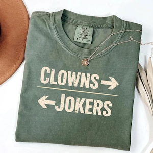 May include: An olive green t-shirt with the words "CLOWNS -> JOKERS" printed in a light tan color. A silver necklace with a small pendant is draped across the shirt. The shirt is folded on a white surface.