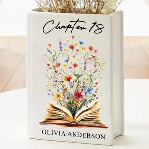 Op de afbeelding: Een witte keramische vaas in de vorm van een boek met de tekst "Chapter 18" en "Olivia Anderson". De vaas heeft een kleurrijk bloemmotief dat uit een open boek komt.