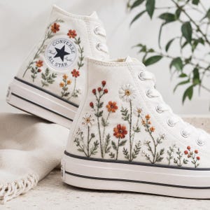 Op de afbeelding: Een paar off-white hoge canvas sneakers met ingewikkeld bloemenborduursel. De schoenen tonen kleurrijke gestikte wilde bloemen in tinten oranje, rood en wit, met groene stengels en bladeren. Het Converse All Star-logo is zichtbaar op de zijkant.