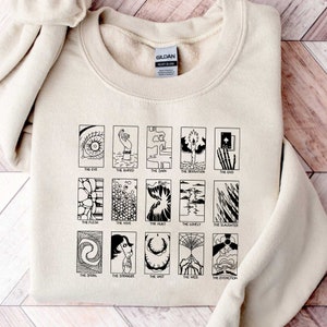 Peut inclure: Un sweat-shirt beige avec un imprimé noir de 15 cartes de tarot. Chaque carte a une illustration unique et un titre, tels que "L'Œil", "L'Enterré", "Les Ténèbres", "La Désolation", "La Fin", "La Chair", "La Ruche", "La Chasse", "Le Solitaire", "Le Massacre", "La Spirale", "L'Étranger", "L'Immensité", "La Toile", et "L'Extinction".