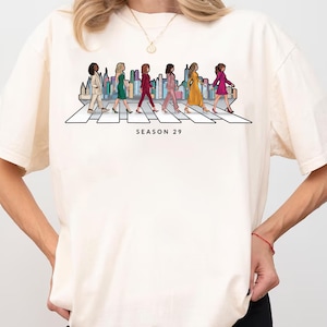 Może przedstawiać: Kremowa koszulka z grafiką przedstawiającą sześć postaci przechodzących przez przejście dla pieszych na tle panoramy miasta. Postacie ubrane są w różne stroje w odcieniach zieleni, czerwieni, różu i żółci. Pod spodem nadruk "SEASON 29".