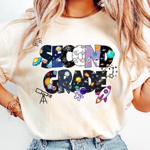 Puede incluir: Camiseta color crema con las palabras "SECOND GRADE" en un diseño colorido con temática espacial. Las letras están llenas de planetas, estrellas, cohetes y un astronauta. También se incluye un telescopio y un OVNI.
