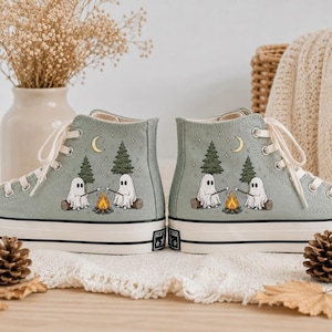 May include: Sage green high-top canvas sneakers featuring a printed graphic of two white ghosts sitting on logs by a campfire, roasting marshmallows under pine trees and a crescent moon. The shoes have white laces and a thick white rubber sole.