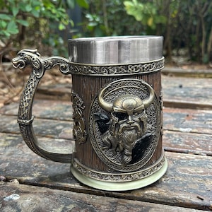 May include: A metal beer stein with a brown wood-like finish. The stein features a raised design of a Viking warrior with a horned helmet, a beard, and ravens. The handle is shaped like a dragon's head.