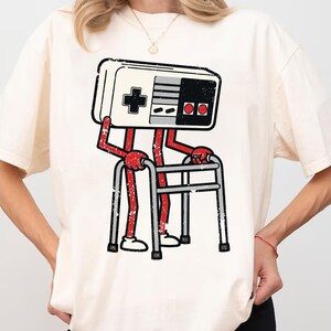 Może przedstawiać: Kremowa koszulka z nadrukiem kontrolera do gier w stylu retro. Kontroler ma czerwone ramiona i nogi, trzymające srebrny chodzik. Na kontrolerze znajduje się czarny krzyż, przyciski i pad kierunkowy. Projekt ma postarzany, vintage wygląd.