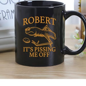 May include: Black ceramic mug with a large handle. The mug features the name "ROBERT" in gold lettering, a cartoon grasshopper, and the phrase "IT'S PISSING ME OFF" in gold.