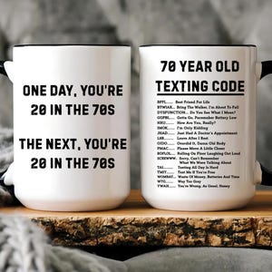 May include: Two white ceramic mugs with black handles and rims. One mug reads "ONE DAY, YOU'RE 20 IN THE 70S. THE NEXT, YOU'RE 20 IN THE 70S." The other mug has "70 YEAR OLD TEXTING CODE" and a list of texting acronyms.