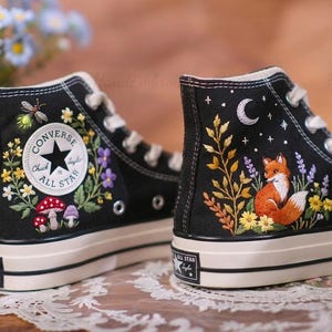 Op de afbeelding: Zwarte high-top Converse Chuck Taylor All Star sneakers met geborduurde bloemen- en bosontwerpen. Eén schoen heeft een libel, bloemen en paddenstoelen. De andere heeft een vos, maan, sterren en bloemen. Witte zolen en veters.