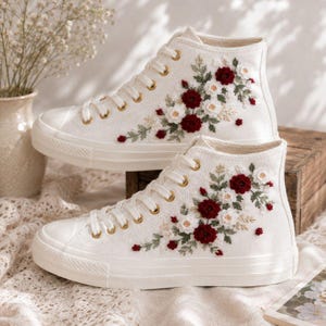 May include: A pair of white high-top canvas sneakers featuring intricate floral embroidery. The design showcases deep red and white blossoms with green leaves and stems. The shoes have white laces, gold-toned eyelets, and a white rubber sole.