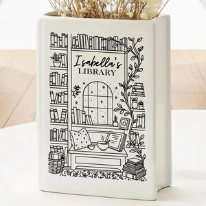 Puede incluir: Jarr&oacute;n de cer&aacute;mica blanco con forma de libro, con una ilustraci&oacute;n en blanco y negro de una biblioteca. El texto "Isabella's LIBRARY" es visible. El jarr&oacute;n contiene flores secas. El dise&ntilde;o incluye estanter&iacute;as y una acogedora zona de lectura.