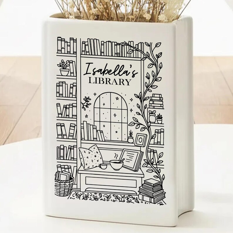 Puede incluir: Jarr&oacute;n de cer&aacute;mica blanco con forma de libro, con una ilustraci&oacute;n en blanco y negro de una biblioteca. El texto "Isabella's LIBRARY" es visible. El jarr&oacute;n contiene flores secas. El dise&ntilde;o incluye estanter&iacute;as y una acogedora zona de lectura.