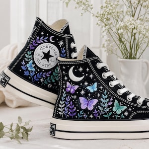 Op de afbeelding: Een paar zwarte hoge sneakers met witte veters en rubberen zolen. De canvas zijkanten zijn versierd met geborduurde paarse, blauwe en turquoise vlinders, bladranken, een witte halve maan en kleine sterretjes. Een rond logoplaatje is zichtbaar.