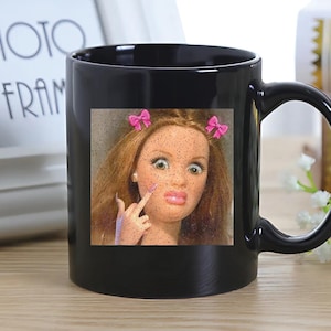 May include: Black ceramic mug featuring a doll image with pink bows and a hand gesture. The doll has red hair, pink lips, and is giving the middle finger. The mug has a curved handle.