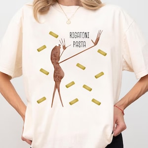 Könnte beinhalten: Cremefarbenes T-Shirt mit einer skurrilen Illustration einer Figur mit ausgestreckten Armen, umgeben von Rigatoni-Nudeln. Die Worte "RIGATONI PASTA" sind in Schwarz über der Figur aufgedruckt. Das Design ist verspielt und hat ein Lebensmittelthema.