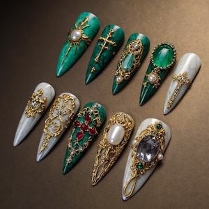 May include: A collection of ten long, pointed artificial nails. The nails feature intricate designs with gold filigree, crosses, pearls, and large gemstones in shades of green, red, and white. The nails are arranged on a brown surface.