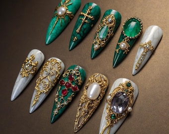 May include: A collection of ten long, pointed artificial nails. The nails feature intricate designs with gold filigree, crosses, pearls, and large gemstones in shades of green, red, and white. The nails are arranged on a brown surface.
