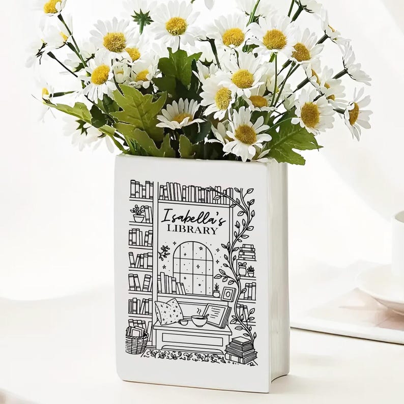 Puede incluir: Un jarr&oacute;n de cer&aacute;mica blanco con forma de libro, lleno de margaritas blancas con centros amarillos y hojas verdes. El jarr&oacute;n est&aacute; decorado con una ilustraci&oacute;n en blanco y negro de una escena de biblioteca, con el texto "Isabella's Library."