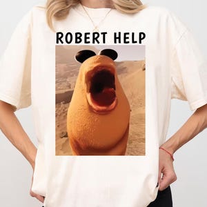 Könnte beinhalten: Cremefarbenes T-Shirt mit einem Meme-Bild einer Zeichentrickfigur mit weit geöffnetem Mund und schwarzer Sonnenbrille. Der Text "ROBERT HELP" ist in schwarzen Blockbuchstaben über dem Bild aufgedruckt. Das Shirt ist ein lässiger Stil.