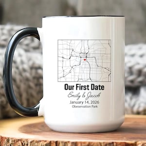May include: White ceramic mug with a black handle and rim. The mug features a map graphic with a red heart, and the text "Our First Date" with names and a date. The mug is on a wooden surface.
