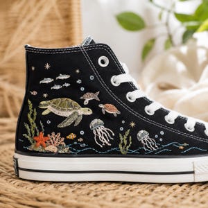 May include: A black high-top canvas sneaker featuring detailed embroidery of an underwater scene. The design includes a green sea turtle, a smaller brown turtle, two jellyfish, a bright orange starfish, coral, seaweed, and small fish.