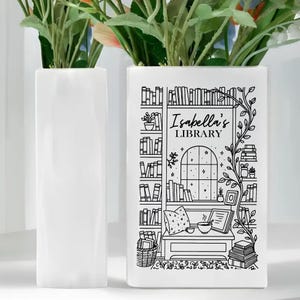 Puede incluir: Dos jarrones rectangulares blancos, uno liso y otro con una ilustraci&oacute;n en blanco y negro. El jarr&oacute;n ilustrado presenta una escena de biblioteca con el texto "Isabella's LIBRARY". Flores verdes y rosas est&aacute;n en los jarrones.