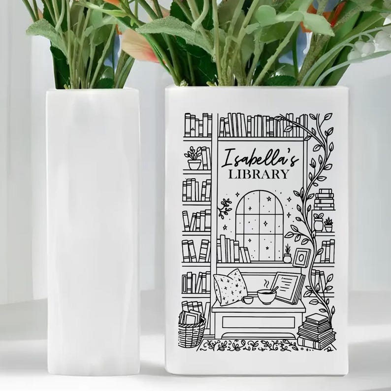 Puede incluir: Dos jarrones rectangulares blancos, uno liso y otro con una ilustraci&oacute;n en blanco y negro. El jarr&oacute;n ilustrado presenta una escena de biblioteca con el texto "Isabella's LIBRARY". Flores verdes y rosas est&aacute;n en los jarrones.