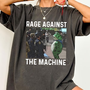 May include: Dark grey t-shirt with the white text "RAGE AGAINST THE MACHINE". The graphic shows a cartoon frog facing police officers. The frog is green and wears a blue scarf. The t-shirt is a casual style.