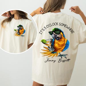 May include: A cream-coloured t-shirt with a vibrant parrot design. The parrot, wearing sunglasses, holds a cocktail. Above the parrot is the text "IT'S 5 O'CLOCK SOMEWHERE", with "Jimmy Buffett" below.