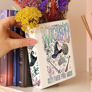Puede incluir: Un jarrón de cerámica blanco con la palabra "WICKED" en letras coloridas, decorado con ilustraciones temáticas de brujas y la frase "CHANGED FOR GOOD". El jarrón contiene un ramo de flores secas.