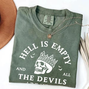 May include: Olive green t-shirt with the phrase "HELL IS EMPTY AND ALL THE DEVILS" printed in white, along with a skull wearing a crown. A gold necklace and a brown hat are also visible.