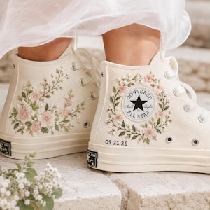 May include: Off-white high-top canvas sneakers featuring delicate floral embroidery in shades of pink and green. The right shoe displays a circular Converse All Star patch surrounded by a floral wreath and the embroidered date 09.21.26.