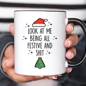 Könnte beinhalten: Weiße Keramiktasse mit schwarzem Henkel und Rand. Die Tasse zeigt einen Weihnachtsmannhut, einen Weihnachtsbaum und den Text "LOOK AT ME BEING ALL FESTIVE AND SHIT" in Schwarz.