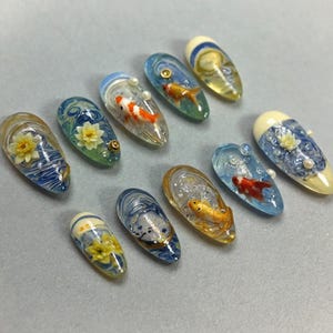 Peut inclure: Collection de dix modèles d'ongles en acrylique. Chaque ongle présente un design unique, notamment des carpes koï, des nénuphars et des motifs tourbillonnants bleus et blancs. Certains ongles ont des accents dorés et de petites perles. Les ongles sont de forme ovale et ont une finition brillante.