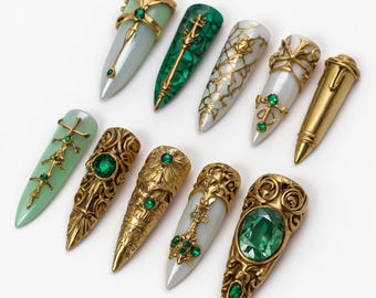 May include: An assortment of ten ornate, pointed nail designs. The nails feature gold and jewel embellishments, including emerald green and pearl-like accents. Some nails have a malachite green or pale green base, while others are white or gold.
