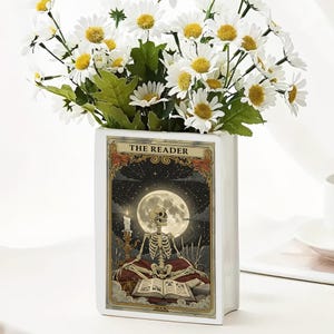 Op de afbeelding: Een witte rechthoekige vaas met een tarotkaartontwerp met een skelet dat onder een volle maan leest. De vaas bevat witte madeliefjes met gele centra en groene bladeren. De tekst "THE READER" staat bovenaan de kaart.