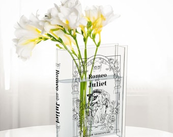 May include: A clear glass vase shaped like a book with the title "Romeo and Juliet" by William Shakespeare printed on the front. The vase is filled with white flowers.