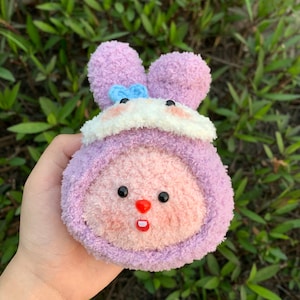 May include: A plush, lavender bunny-shaped toy with a white and blue accent. The toy has black eyes, a red nose, and pink cheeks. The bunny is made of soft, fuzzy material.