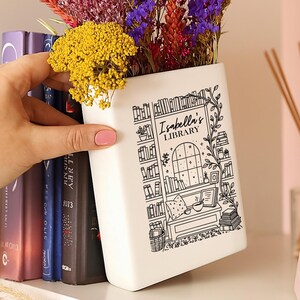 Puede incluir: Un jarr&oacute;n rectangular blanco con una ilustraci&oacute;n negra de una escena de biblioteca, que incluye estanter&iacute;as y una ventana, y el texto "Isabella's LIBRARY". El jarr&oacute;n contiene un ramo de flores secas de colores. El jarr&oacute;n es sostenido por una persona.