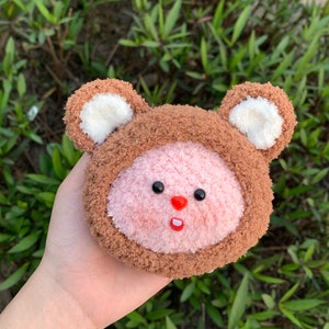 May include: A brown and pink plush toy shaped like a bear with white ears. The bear has a pink face with black eyes and a red nose. The bear's mouth is open and has a white tooth showing.