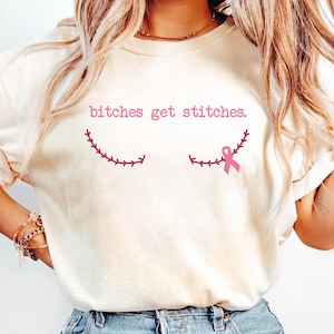 May include: A cream-coloured t-shirt featuring the pink text "bitches get stitches". Below the text are two pink stitched designs, with a pink ribbon on the right. The shirt is a casual style.