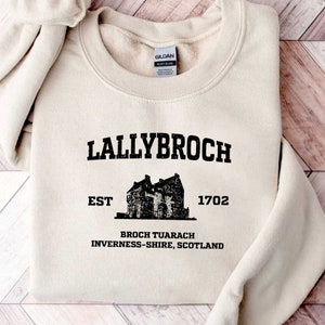 Könnte beinhalten: Ein beiger Pullover mit einem schwarzen grafischen Druck eines Schlosses und dem Text "Lallybroch Est 1702 Broch Tuarach Inverness-shire, Schottland".