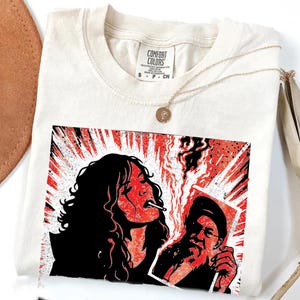 May include: Cream-coloured Comfort Colors t-shirt with a red and black graphic of a woman smoking a cigarette and a man holding a photo. A gold necklace with a pendant is draped across the shirt.