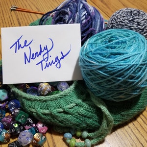 Può includere: Una borsa di maglia verde piena di dadi colorati e un biglietto che dice "The Nerdy Things". Ci sono anche tre gomitoli di lana sullo sfondo, uno blu, uno viola e uno bianco e nero.
